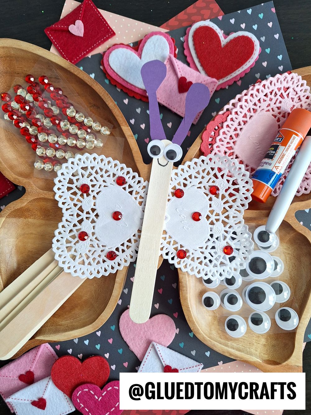 Valentine’s Day butterfly craft made from a popsicle stick, heart doilies, googly eyes, felt hearts, and red gems—perfect for those seeking fun Valentine’s Day craft ideas for kids. Surrounded by extra craft supplies and decorations on a table.