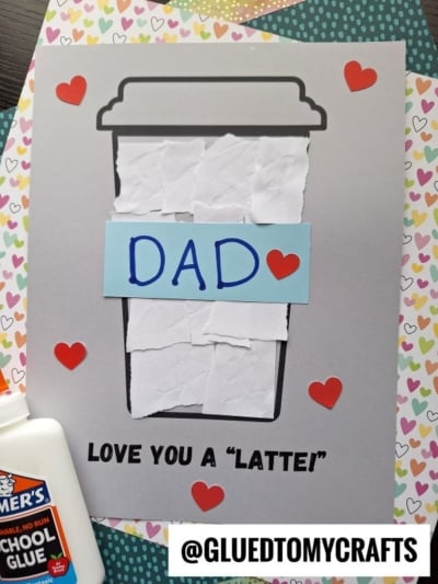 A handmade card showcases a torn paper latte craft, with a coffee cup made from white paper, DAD and a red heart on the sleeve. Red heart stickers decorate the card. Text below reads, LOVE YOU A LATTE! and a glue bottle sits nearby.