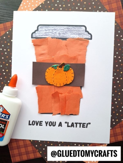 A handmade coffee cup card features a charming torn paper latte craft, showcasing torn orange paper, a brown band with a pumpkin, and text that says LOVE YOU A LATTE. A bottle of Elmer’s glue sits nearby on a polka dot background.