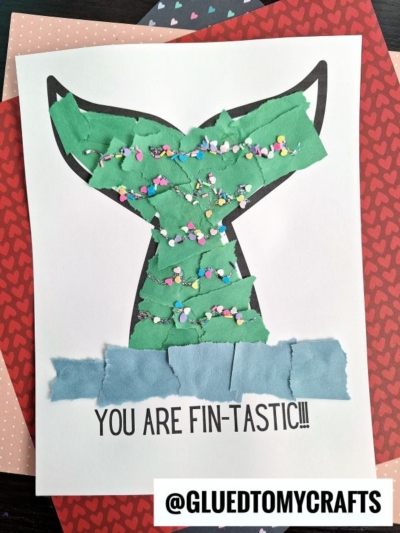 A handmade torn paper mermaid tail craft with green and blue pieces, decorated with colorful confetti and the text YOU ARE FIN-TASTIC!!! at the bottom. The handle @GLUEDTOMYCRAFTS appears in a box.