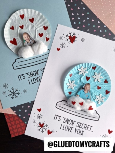 Two handmade cupcake liner snow globe cards feature a child’s photo inside each globe, accented with red hearts, white snowflakes, and cotton snow. “It’s ‘snow’ secret I love you!” appears on both cards. @GLUEDTOMYCRAFTS at the bottom.
