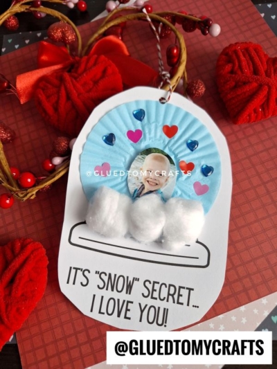A Valentine’s Day craft featuring a child’s photo inside a cupcake liner snow globe, with heart stickers, cotton ball snow, red hearts, and the sweet message: “It’s ‘snow’ secret... I love you!”.