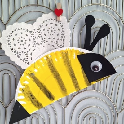 A bee craft made from a painted paper plate with yellow and black stripes, a heart-shaped doily for wings, and a googly eye—perfect for valentine's day craft ideas for kids. Displayed on a textured background. @GLUEDTOMYCRAFTS is written at the bottom.