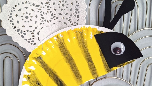 A bee craft made from a painted paper plate with yellow and black stripes, a heart-shaped doily for wings, and a googly eye—perfect for valentine's day craft ideas for kids. Displayed on a textured background. @GLUEDTOMYCRAFTS is written at the bottom.