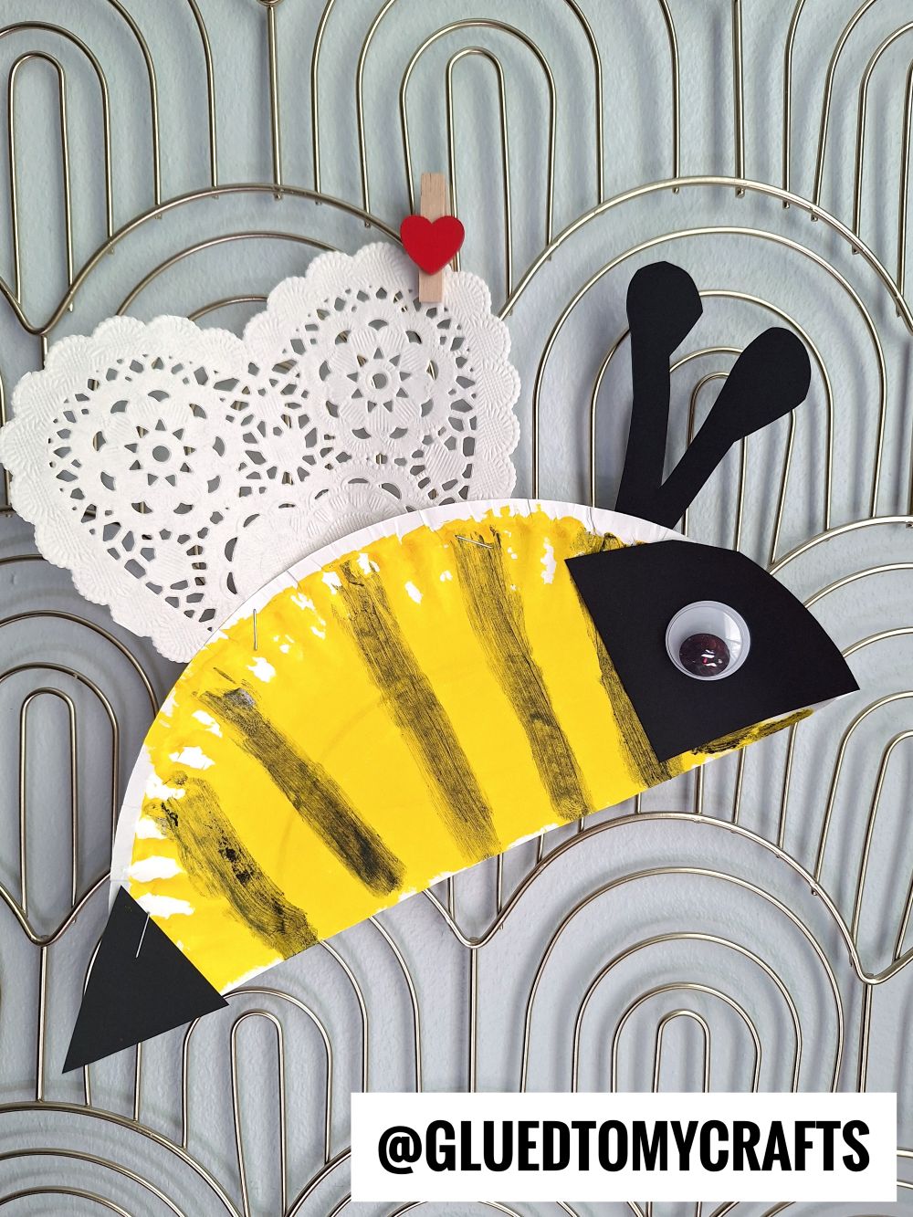 A bee craft made from a painted paper plate with yellow and black stripes, a heart-shaped doily for wings, and a googly eye—perfect for valentine's day craft ideas for kids. Displayed on a textured background. @GLUEDTOMYCRAFTS is written at the bottom.