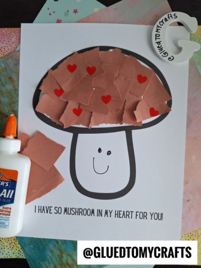 A torn paper mushroom craft with a smiling face is decorated with red heart stickers. A bottle of glue and a round Glued To My Crafts label are nearby. The text says, I have so mushroom in my heart for you!.