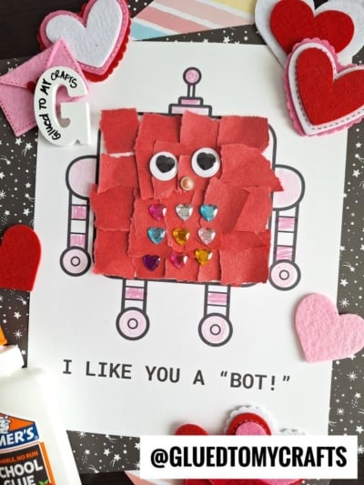 A torn paper robot made of red scraps and googly eyes is glued to a printed outline. Colorful heart stickers decorate its body. Felt hearts, glue, and craft supplies surround the project. Text reads I LIKE YOU A BOT!.