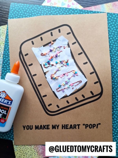 A handmade craft card features a torn paper pastry shaped like a rectangular toaster treat, decorated with glued-on beads and painted lines, next to a bottle of Elmer’s glue. Text reads, You make my heart pop!.