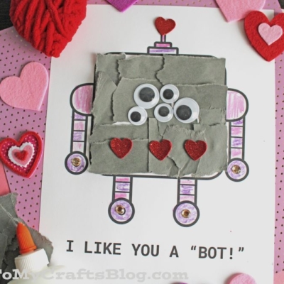 A handmade torn paper robot Valentine craft on white paper reads I LIKE YOU A BOT! The robot features googly eyes, red glitter hearts, gray textured material, and is surrounded by pink and red felt hearts with school glue.