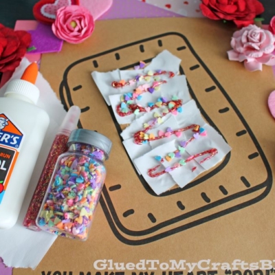 A craft project decorated with torn paper pastry faux sprinkle pop-tarts, felt hearts, paper flowers, a glue bottle, and colorful sprinkles on a pink polka-dot background.