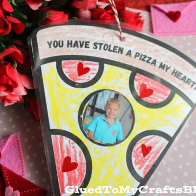 A slice-shaped card decorated like pizza with hearts, featuring a child's photo as a pizza photo ornament in the center. Surrounded by red felt envelopes and hearts. Text reads, YOU HAVE STOLEN A PIZZA MY HEART!