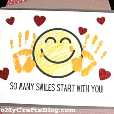 A handmade card featuring a cheerful handprint smile with a large yellow smiley face, two yellow handprints on each side, red glitter hearts, and the text SO MANY SMILES START WITH YOU! on crisp white paper.