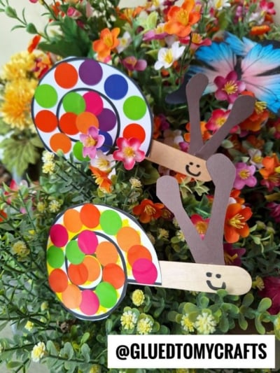 Two polka dot snail crafts made from paper and colorful dot stickers sit among vibrant flowers. Each snail has a smiling face and antennae crafted from brown paper and sticks. @GLUEDTOMYCRAFTS is written at the bottom.
