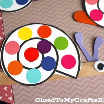 A polka dot snail craft made from paper and a popsicle stick, featuring a spiral shell with rainbow colored dots