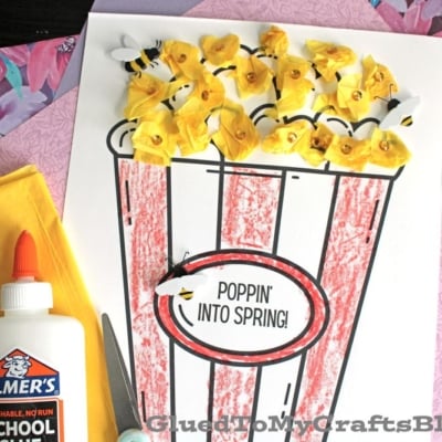 A spring-themed craft featuring a popcorn bucket outlined in red, filled with crumpled yellow tissue paper and paper bees. Scissors, glue, and paper are nearby.