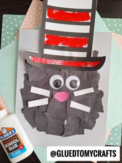 A handmade torn paper cat craft inspired by the Cat in the Hat, featuring black paper for the face, large googly eyes, white whiskers, a pink nose, and a red-and-white striped hat. Elmer’s glue is on the side.
