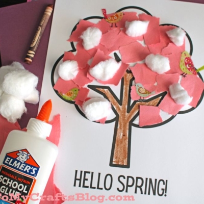 A torn paper cherry blossom tree craft with pink paper and cotton balls glued onto a tree outline. “HELLO SPRING!” is written below, with Elmer’s glue and paper scraps nearby for this fun spring project.