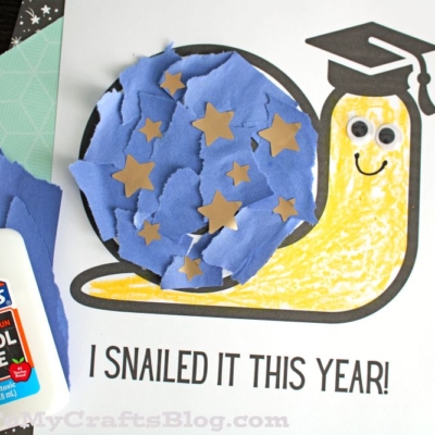 A torn paper snail craft shows a smiling snail with a blue, star-decorated shell and a graduation cap. Googly eyes add charm above the “I SNAILED IT THIS YEAR!” text. Glue and crafting materials are scattered nearby.