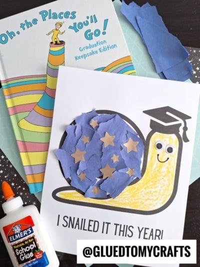 A children's craft project features a torn paper snail with a graduation cap on a worksheet reading "I snailed it this year!" The snail's shell is decorated with blue paper and gold stars. A bottle of glue and a Dr. Seuss book are nearby.