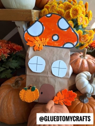 A charming paper bag mushroom house, complete with an orange cap, white windows, brown door, felt leaves, and a pumpkin cutout. Surrounded by pumpkins and orange flowers. Text: @GLUEDTOMYCRAFTS.