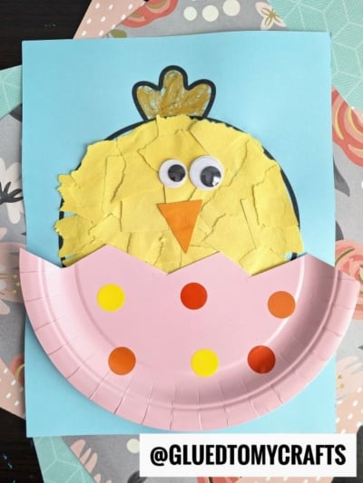 A torn paper Easter chick craft featuring yellow paper, googly eyes, an orange beak, and a pink plate with colorful dots on a blue background. The cute chick looks like it’s hatching from its egg!.