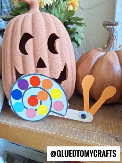 A colorful polka dot snail craft sits in front of a smiling jack-o’-lantern and an orange pumpkin. The snail’s shell is decorated with bright circular stickers. Text at the bottom reads “@GLUEDTOMYCRAFTS.”.