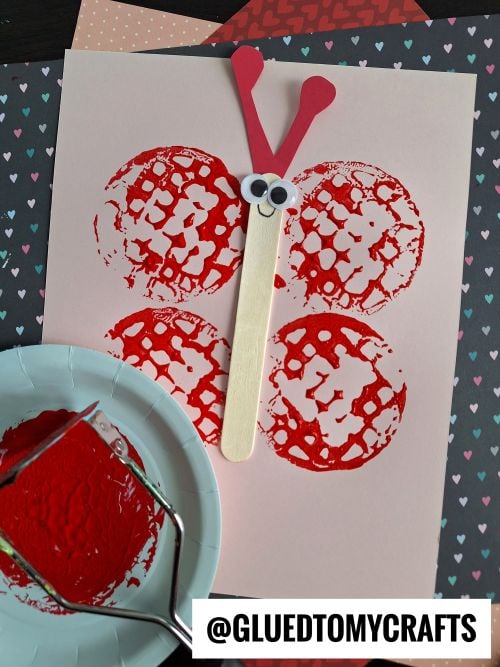 A butterfly potato masher stamped craft made with four red paint-printed circles for wings, a popsicle stick body, googly eyes, and red antennae, displayed on pink paper with craft supplies nearby.