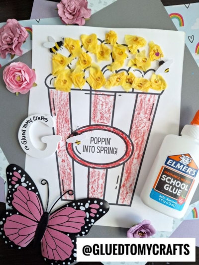 A poppin' into spring-themed popcorn craft features a red-striped box with yellow paper flowers, tiny bees, a cheerful “Poppin’ into Spring!” sign, glue, a paper butterfly, and decorative flowers on a colorful surface.