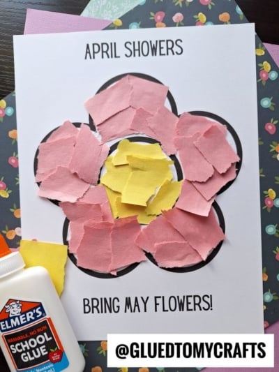A torn paper flower outline is filled with pink and yellow scraps. Text reads April showers bring May flowers! A bottle of Elmer’s School Glue sits beside the project on a floral background.