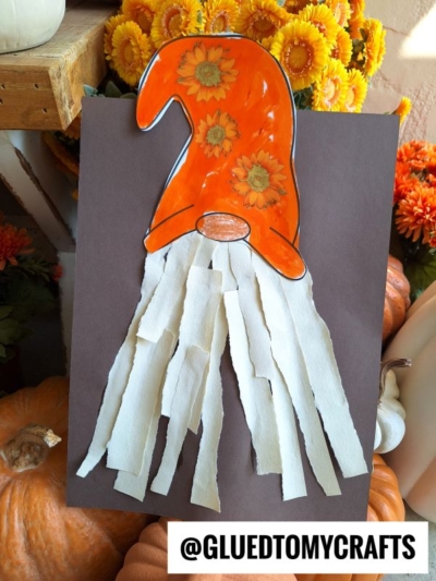 A fall torn paper gnome craft featuring an orange sunflower hat and a long white paper strip beard on dark paper. Surrounded by pumpkins and orange flowers, this charming scene has @GLUEDTOMYCRAFTS text at the bottom.