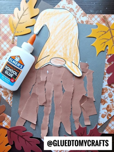 A cozy torn paper gnome craft with a brown beard, yellow hat, and gray background. Surrounded by autumn leaves, plaid paper, and Elmer’s glue for a perfect fall touch. @GLUEDTOMYCRAFTS at the bottom.
