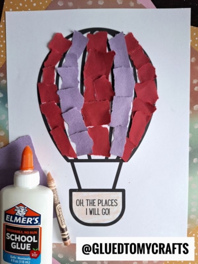 A vibrant torn paper hot air balloon craft made with pieces of red and purple paper on a white sheet, next to a bottle of Elmer’s glue and a brown crayon. The balloon reads, OH, THE PLACES I WILL GO!.