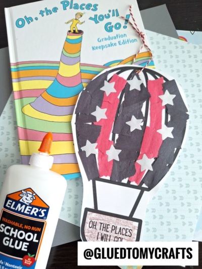 A handmade torn paper hot air balloon craft, decorated with star and stripe patterns, sits in front of the book “Oh, the Places You’ll Go!” alongside a bottle of Elmer’s school glue on a patterned surface.