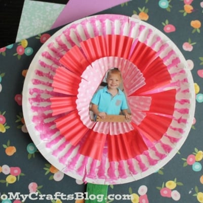 A Paper Plate Flower Photo Frame made with cupcake liner flowers and a child’s photo in the center is displayed on floral-patterned paper.