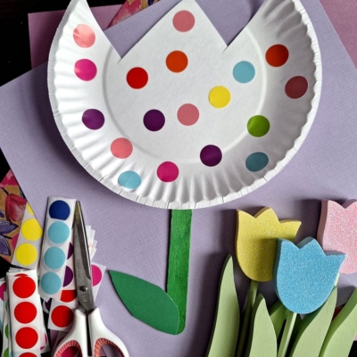 A paper plate decorated with colorful dot stickers to look like a tulip, alongside craft scissors, coding label crafts supplies, paper leaves, and tulip cutouts on a purple background. Text reads @GLUEDTOMYCRAFTS.