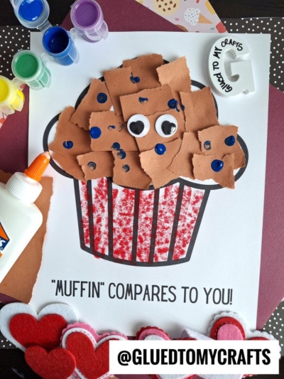 A kids craft project featuring a torn paper muffin made of brown pieces, googly eyes, and blue dots on a white sheet with the caption Muffin compares to you! surrounded by paint, glue, and craft supplies.