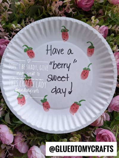 A white paper plate decorated with red strawberry drawings, pink and green flowers, and a handwritten message: “Have a ‘berry’ sweet day!”—a charming example of thumbprint crafts. Image credit: @GLUEDTOMYCRAFTS.
