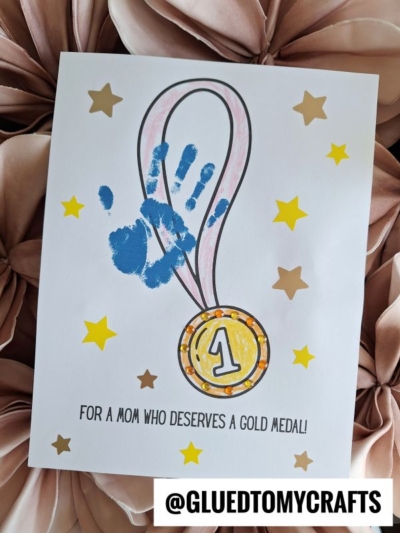 A card featuring a handprint gold medal design with a blue handprint and shimmering gold medal, surrounded by stars. Text reads, For a mom who deserves a gold medal! @GLUEDTOMYCRAFTS is at the bottom.