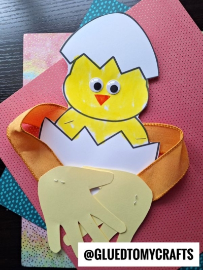 A handmade craft featuring a yellow paper handprint chick hug with googly eyes hatching from a white eggshell, orange ribbon, and colorful paper backgrounds. @GLUEDTOMYCRAFTS appears at the bottom.