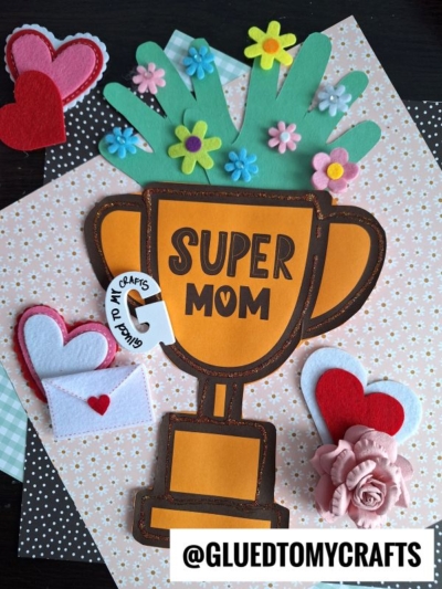 A handmade handprint Mother's Day trophy with Super Mom text, decorated with felt flowers, surrounded by felt hearts, a paper envelope, and a pink paper rose, all arranged on patterned paper. @GLUEDTOMYCRAFTS is at the bottom.