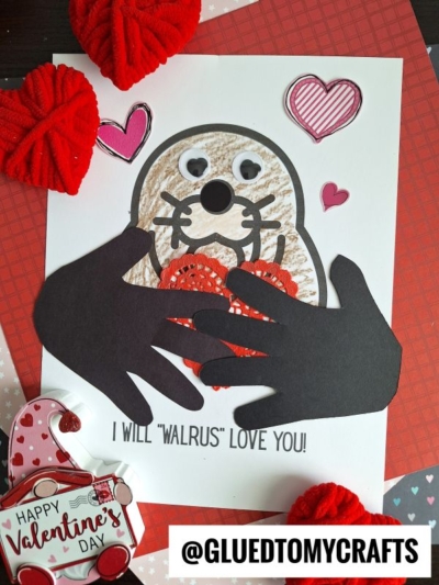 A handmade Valentines Day card features a handprint walrus drawing with googly eyes, a heart-shaped nose, red crocheted hearts, and black cut-out hands. The card reads, I will walrus love you! Hearts and yarn are nearby.