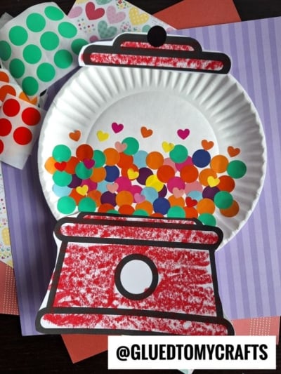 A paper plate gumball machine craft colored with red crayon, decorated with colorful round and heart-shaped stickers. Sheets of unused stickers and patterned papers are displayed in the background.