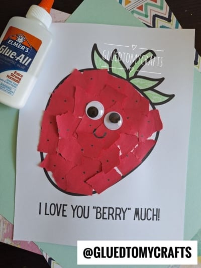 A torn paper strawberry craft made with red paper pieces on a printed outline, topped with googly eyes and a smile. Elmer’s Glue and patterned paper are nearby, with text reading I LOVE YOU BERRY MUCH!.