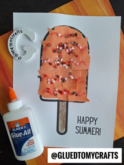 A fun summer craft project features a torn paper popsicle decorated with orange tissue, pink and red heart sprinkles, and glue. A bottle of Elmer’s glue sits nearby. HAPPY SUMMER! and @GLUEDTOMYCRAFTS are printed on the paper.