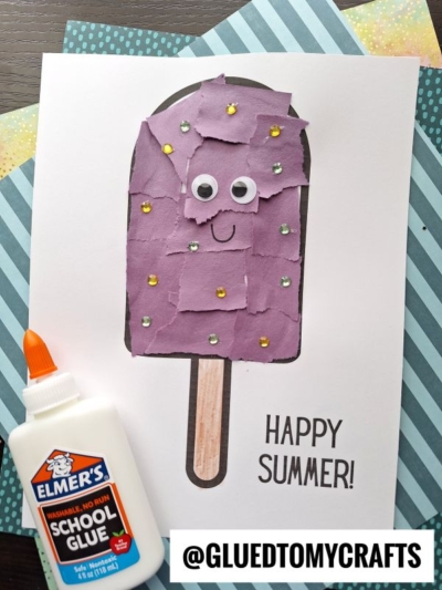 A torn paper popsicle craft made from purple scraps, with googly eyes and gem stickers on white paper saying Happy Summer! A bottle of Elmer’s school glue lies beside it. Background features striped and yellow patterned papers.