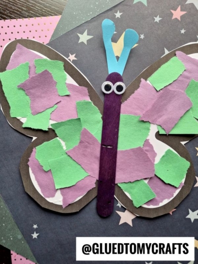 A vibrant torn paper butterfly craft featuring green and purple torn paper wings, a purple stick body, googly eyes, and blue antennae set against a dark star-patterned background. Text: @GLUEDTOMYCRAFTS.