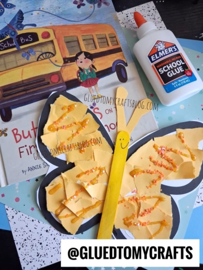 A handmade yellow torn paper butterfly craft with a popsicle stick body sits on top of a children’s book, next to a bottle of Elmer’s glue and scattered sheets of colored paper. Text reads @GLUEDTOMYCRAFTS.