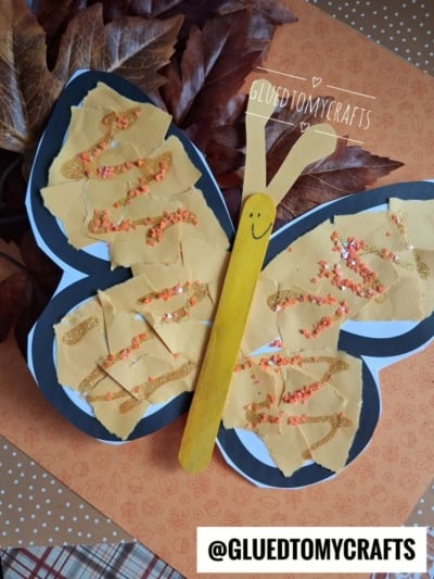 A torn paper butterfly craft made with vibrant yellow paper pieces, glitter accents, and a yellow popsicle stick for the body, placed on an orange leaf background. The handle is decorated with a cheerful smiling face.