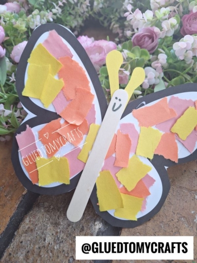 A handmade torn paper butterfly craft with a black outline, white wings decorated with yellow, orange, and pink paper pieces, and a stick body, surrounded by flowers. @GLUEDTOMYCRAFTS is written at the bottom.