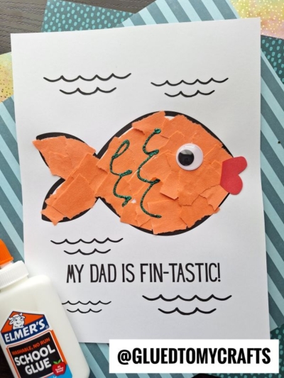 A handmade Fathers Day card for a fin-tastic dad features a fish made of torn orange paper, a googly eye, red paper lips, and wavy green glitter. "MY DAD IS FIN-TASTIC!" is written on the front. Elmers glue sits beside the card.